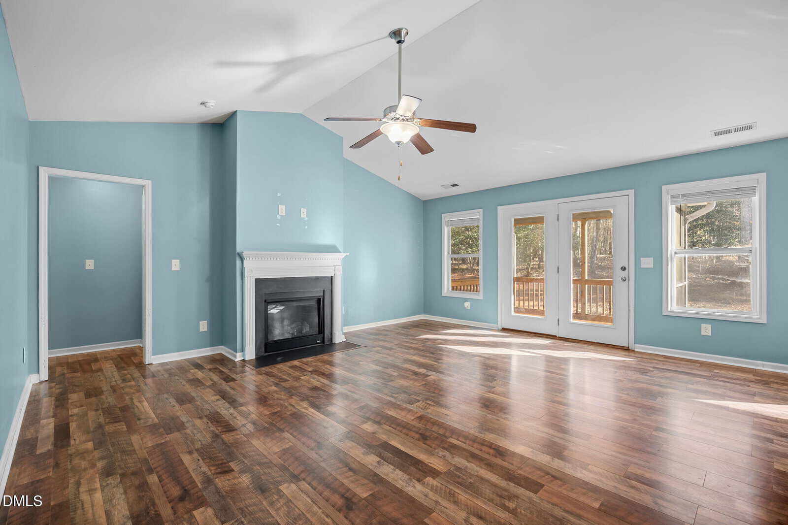 Spacious living room with blue walls, wood flooring, a ceiling fan, white fireplace, and large windows and doors leading to a deck outside. Natural light fills the room.