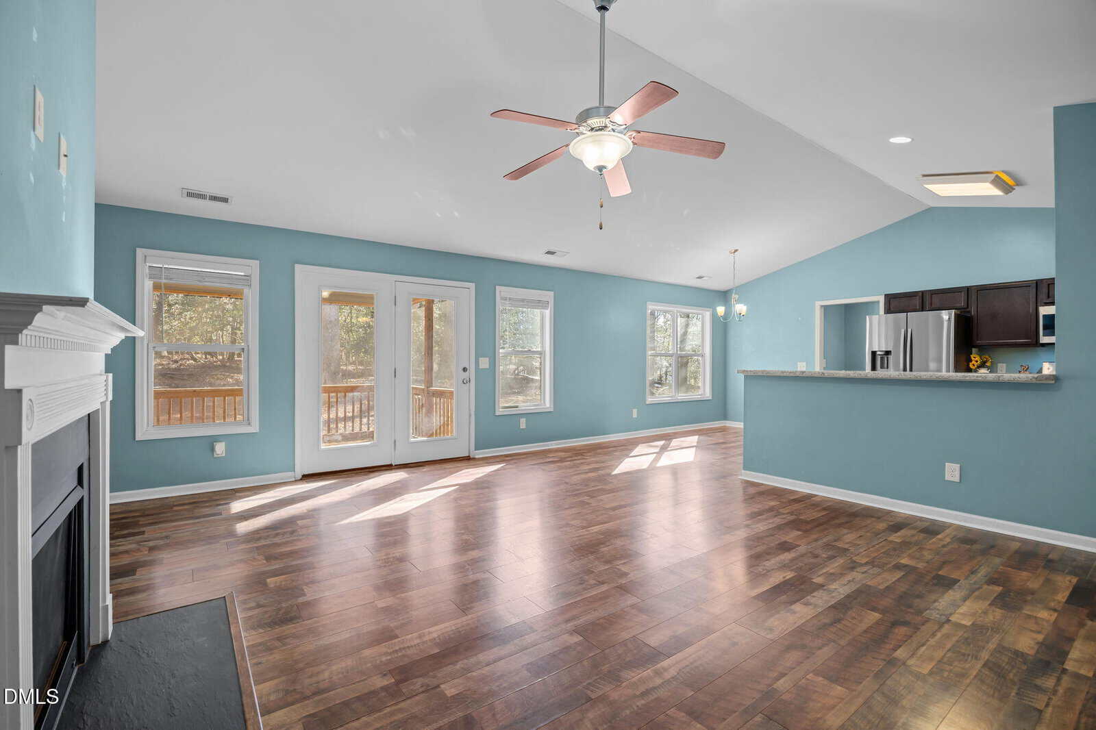 Spacious open-plan living area with wood floors, blue walls, ceiling fan, fireplace, large windows, French doors to a deck, and a modern kitchen with stainless steel appliances and a breakfast bar.