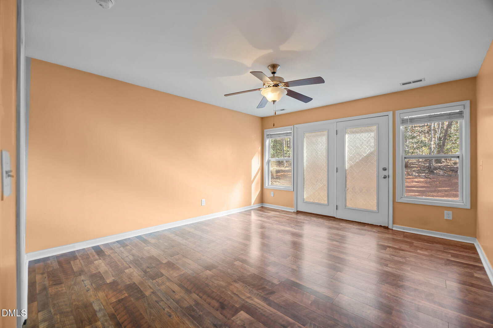 Empty room with peach-colored walls, wood flooring, ceiling fan with light, two windows, and double glass doors leading outside. Natural light enters from the windows and doors. The room appears clean and spacious.