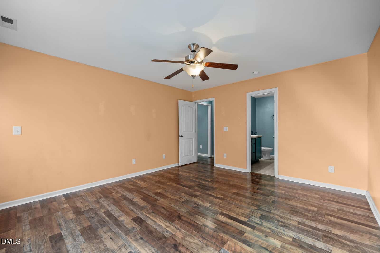 Empty room with hardwood floors, peach-colored walls, a ceiling fan with lights, and two doorways—one open to a bathroom with blue walls and the other to a closet or hallway. No furniture is present.