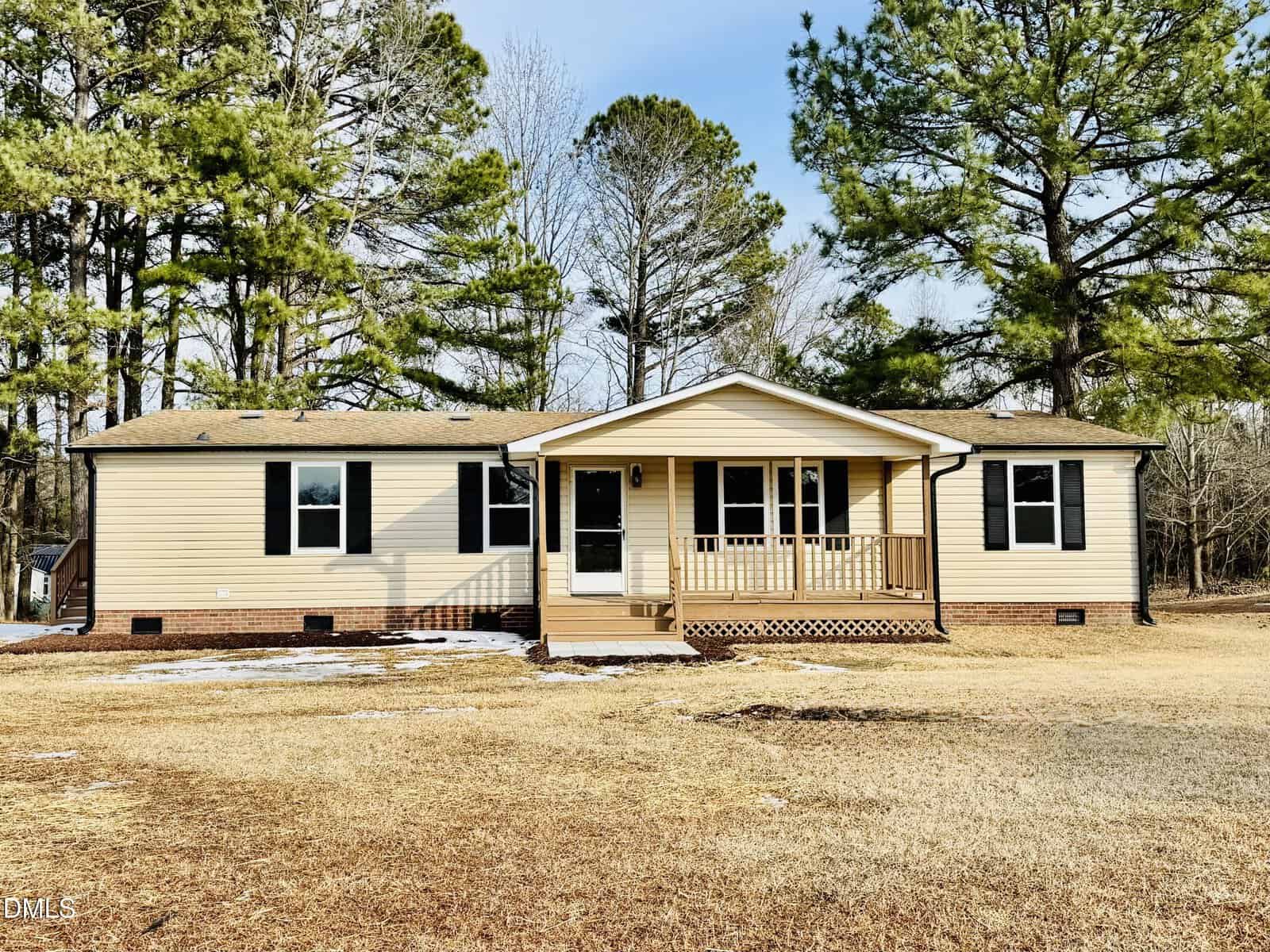 A beige, single-story house with black shutters, a small covered porch, and a brick foundation sits on a large, grassy lot with tall pine trees in the background under a clear sky.