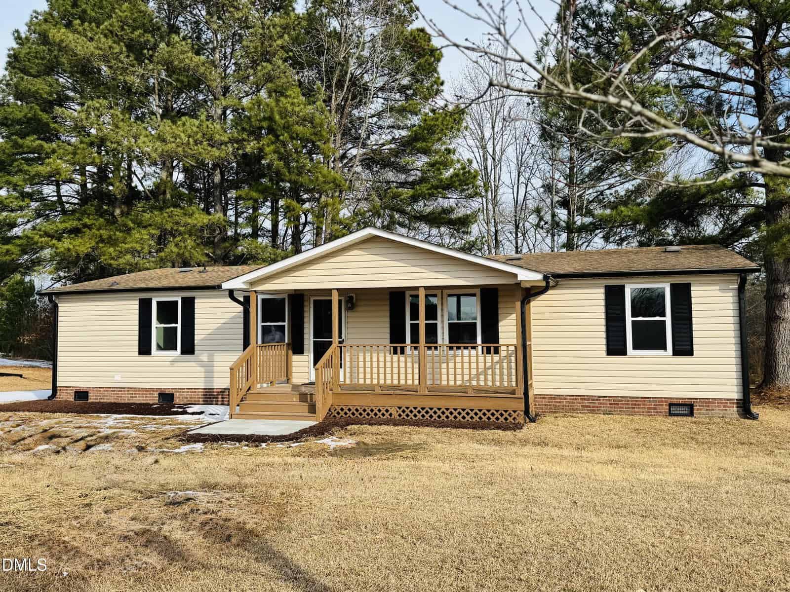 Single-story beige house with black shutters and a covered front porch, surrounded by tall pine trees and dry grass. There is a small patch of snow near the porch and a clear blue sky overhead.