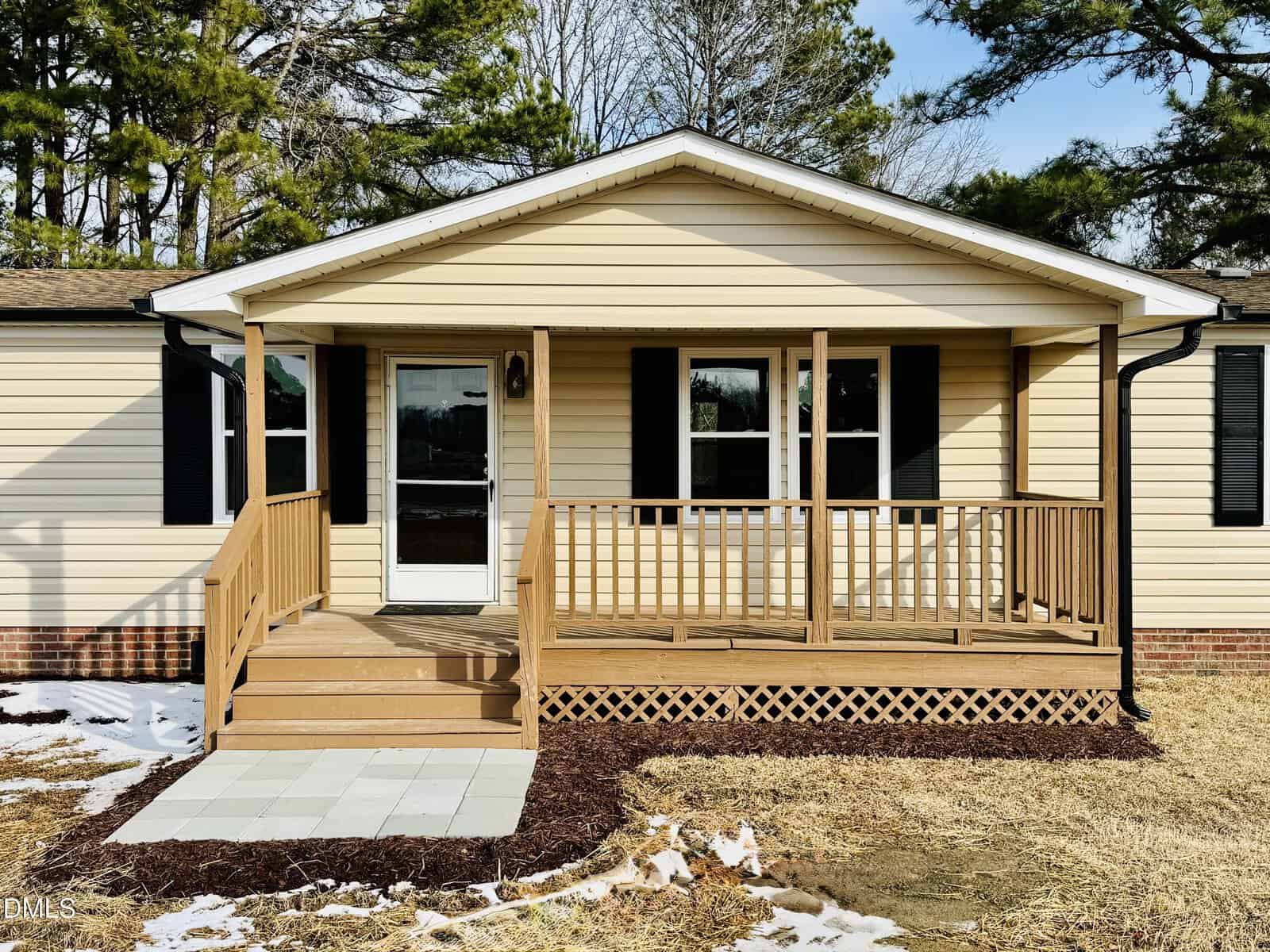 A small beige house with black shutters and a covered front porch featuring wooden railings and steps. There is a tiled path leading to the porch, with patches of snow and trees in the background.