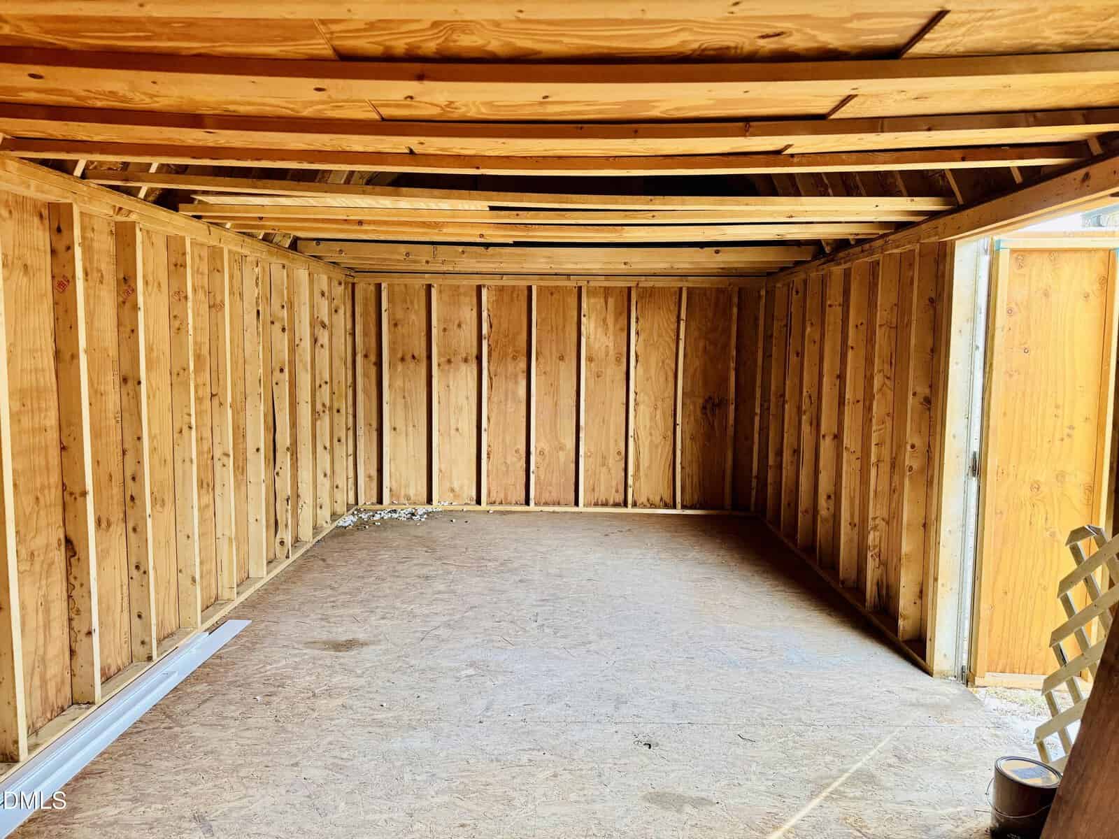An empty wooden shed with exposed framing, plywood floor, and open ceiling beams. Sunlight enters through a partially open door on the right, illuminating the interior.