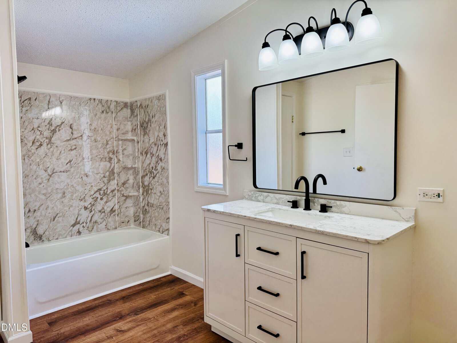 A modern bathroom with a white vanity, marble countertop, black fixtures, a large mirror with lights, wood flooring, and a bathtub with marble-patterned walls. A frosted window is above the sink.