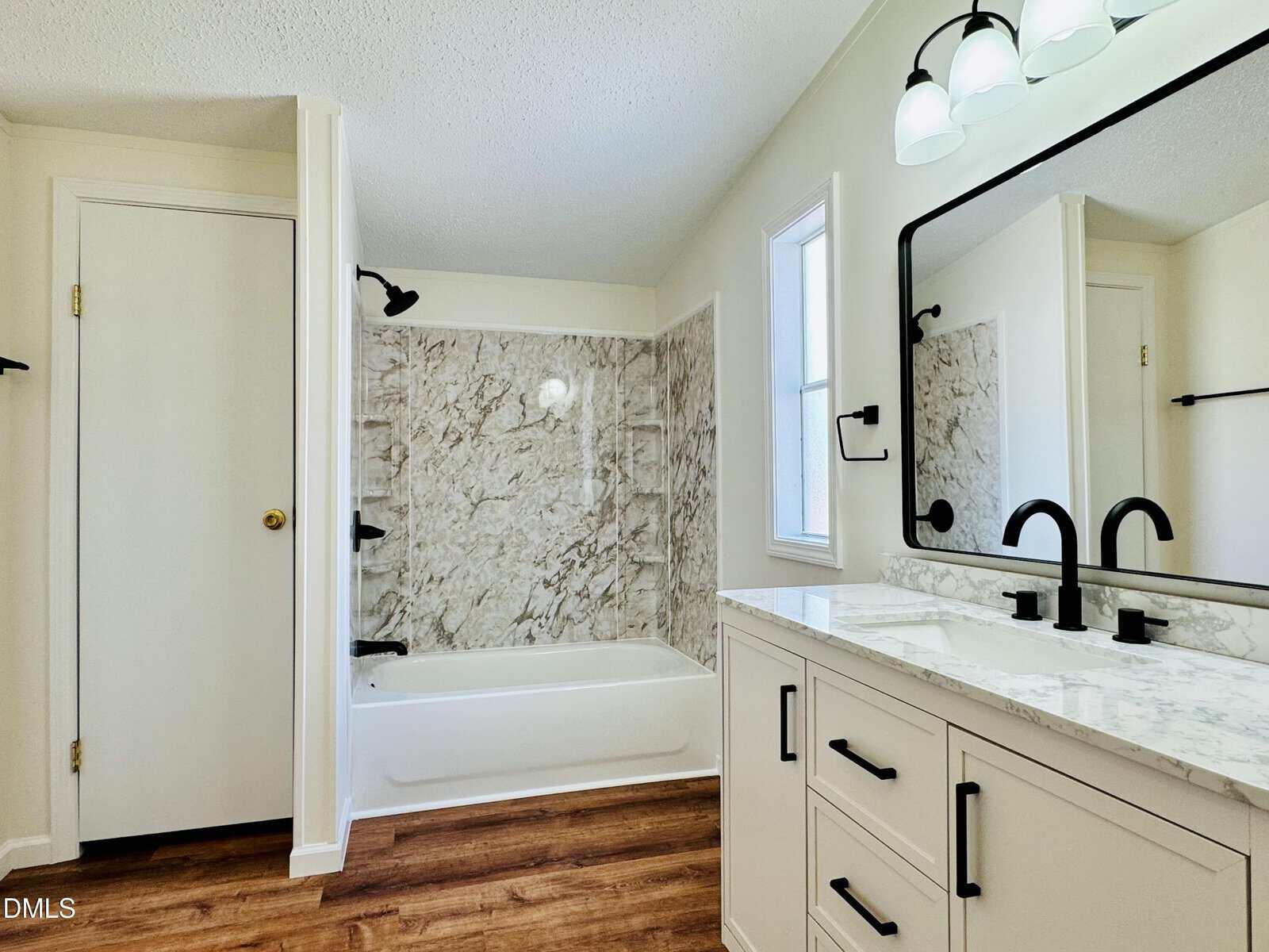 Modern bathroom with a white bathtub and marble-patterned shower surround, white vanity with marble countertop, black fixtures, large mirror, wood-look flooring, and a door to the left.