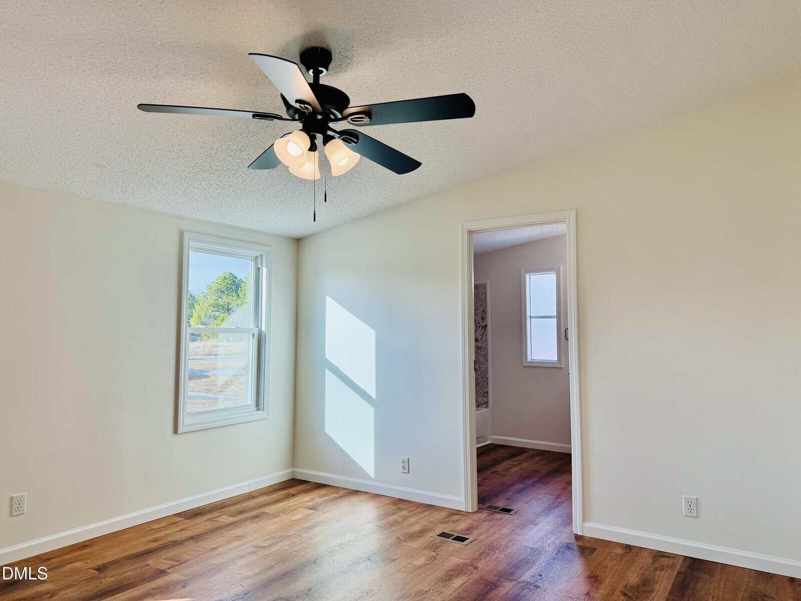 Bright, empty room with white walls, a window letting in sunlight, wood flooring, a black ceiling fan with lights, and an open doorway leading to another room with a window.