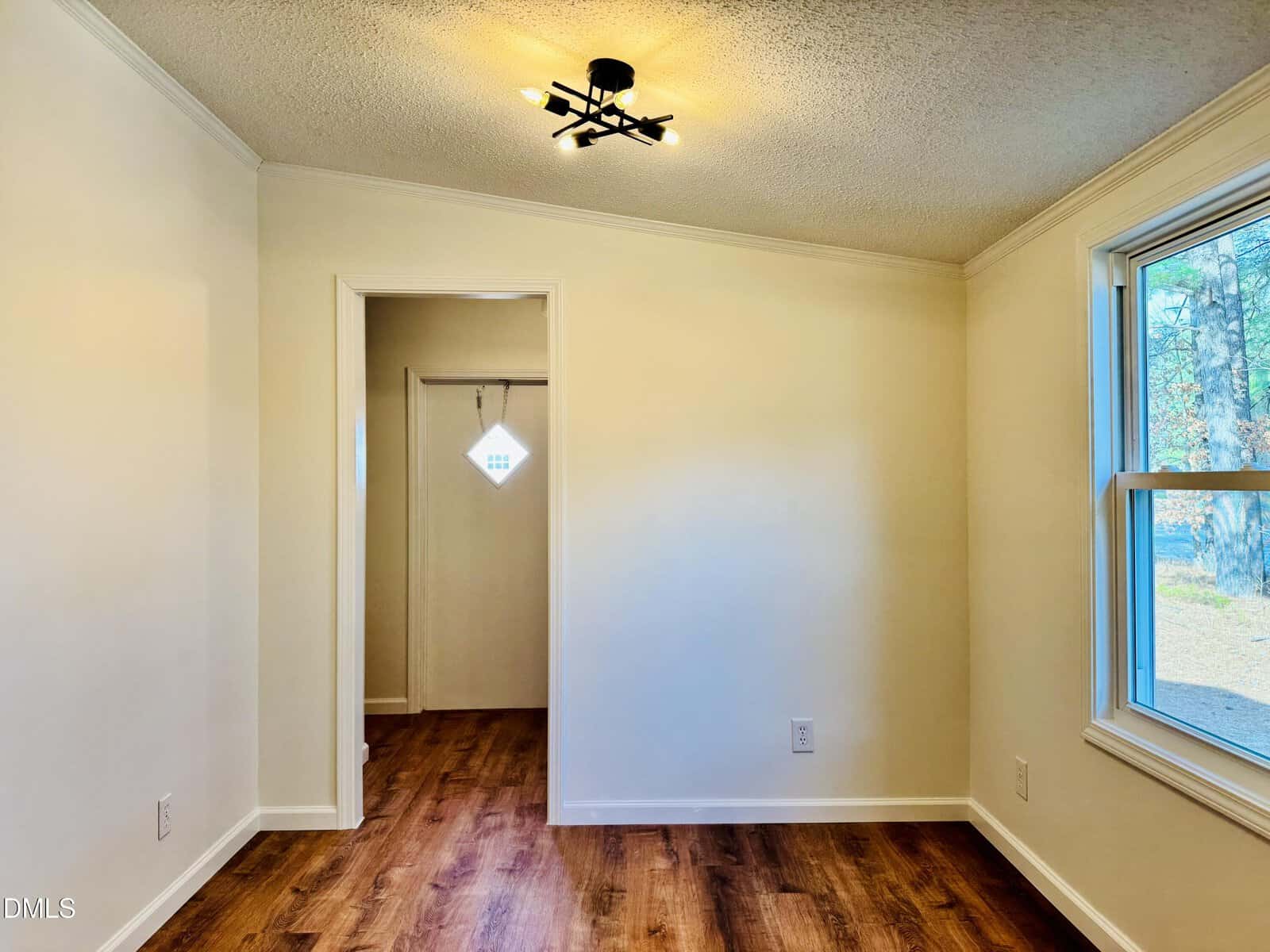 A small, empty room with cream-colored walls, a modern black ceiling light, wood flooring, a window on the right, and an open doorway leading to an entryway with a frosted glass door.