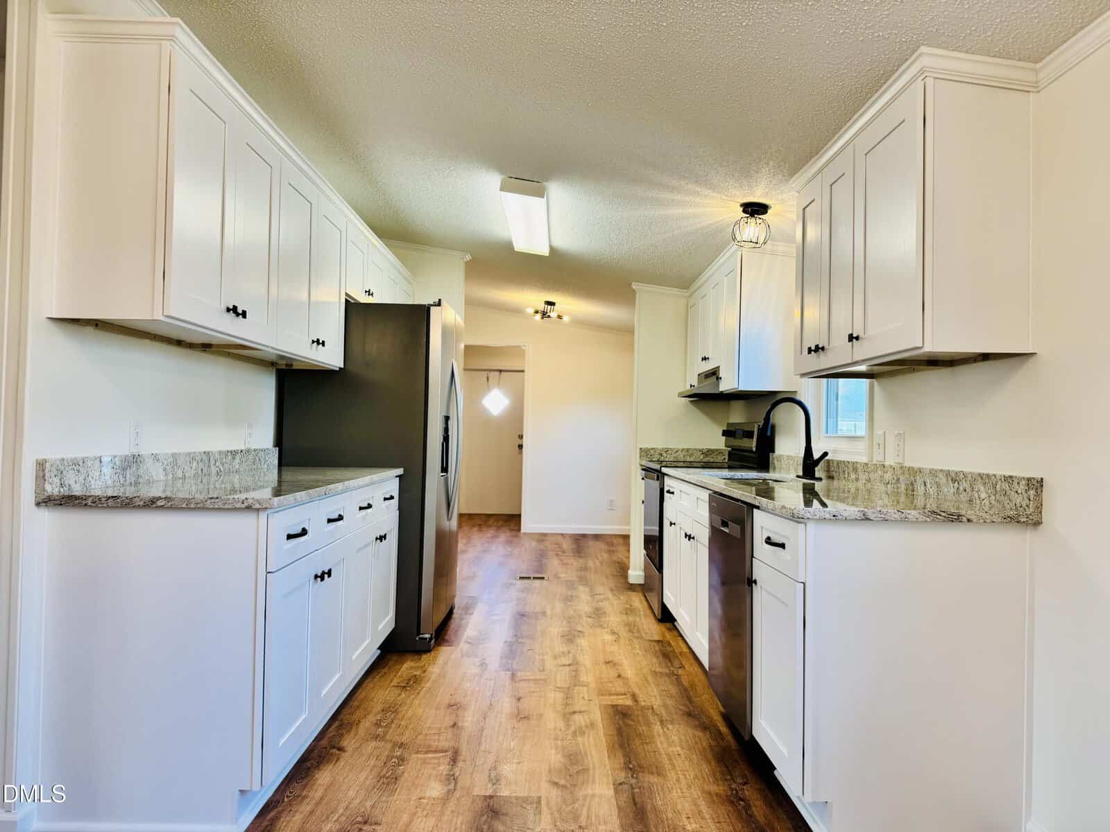 Modern galley kitchen with white cabinets, stainless steel appliances, granite countertops, and wood flooring. A window above the sink and a door with a diamond-shaped window are visible at the far end.