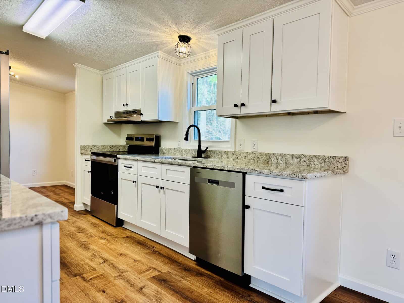 Modern kitchen with white cabinets, stainless steel appliances, a black faucet, granite countertops, and wood flooring. A window above the sink lets in natural light. The space is clean and well-lit.