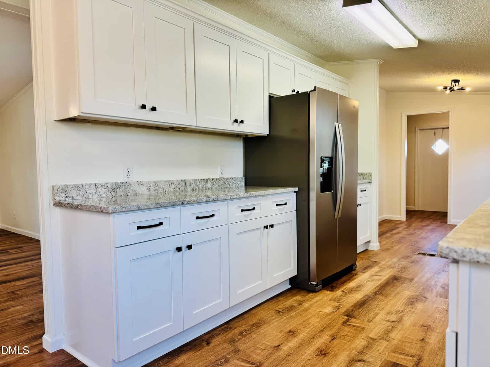 Modern kitchen with white cabinets, stainless steel refrigerator, granite countertops, and wood flooring. Bright lighting and clean, minimalist design.