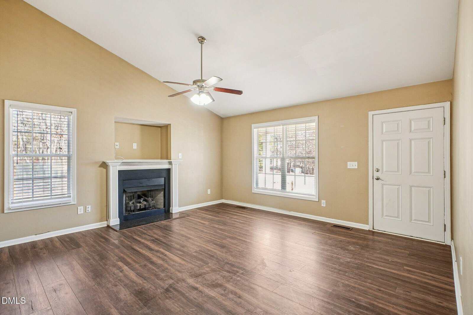A bright, empty living room with tan walls, dark wood floors, a white front door, two windows, a ceiling fan, and a black fireplace with a white mantel.