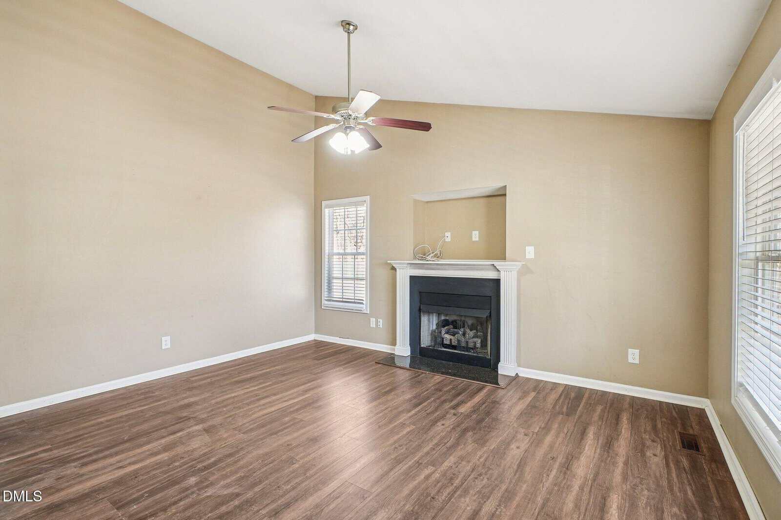 A bright, empty living room with beige walls, wood flooring, a ceiling fan, white-trimmed windows, and a fireplace with a white mantel set into a recessed nook.