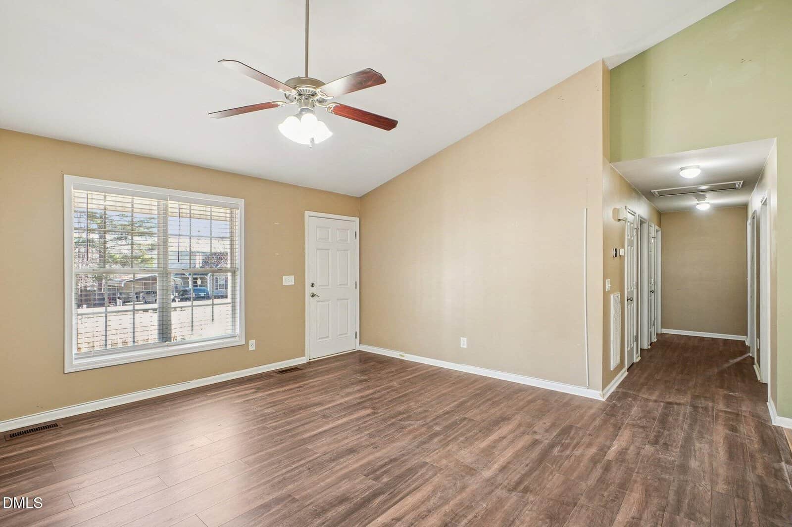 A bright, empty room with beige walls, wood flooring, a ceiling fan, large window, white door, and hallway with recessed lighting leading to other rooms.