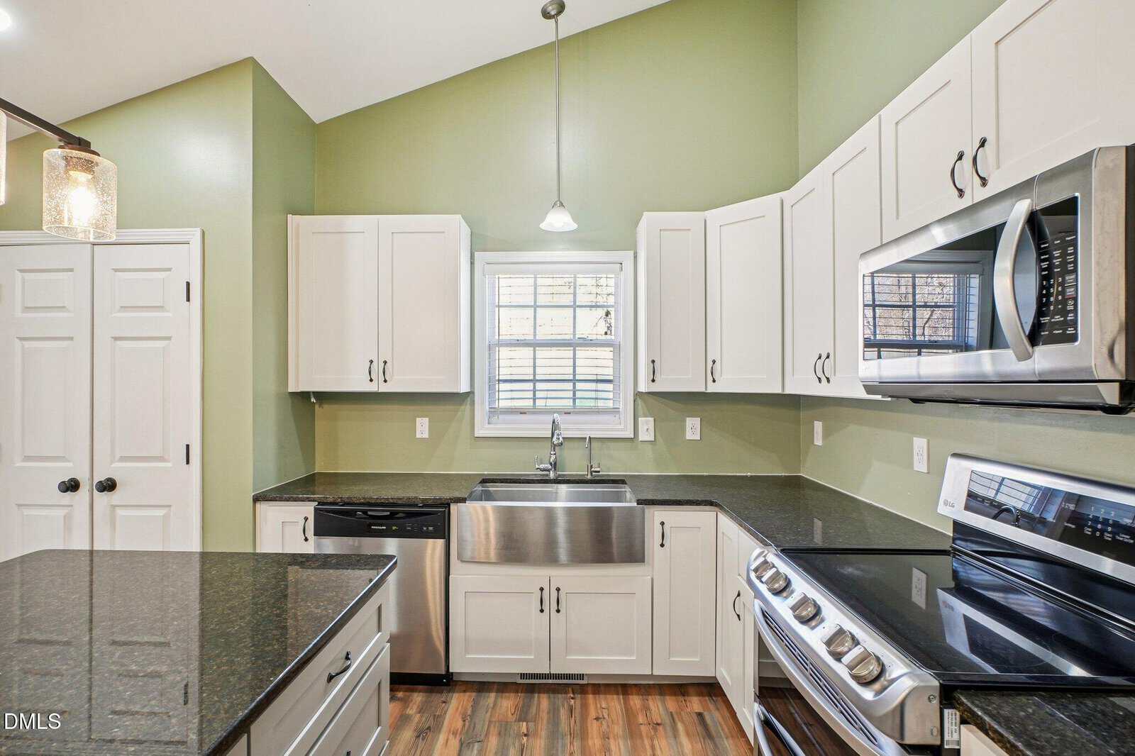 Modern kitchen with green walls, white cabinets, stainless steel appliances, large farmhouse sink under a window, black granite countertops, and wood flooring. A kitchen island is partially visible on the left.
