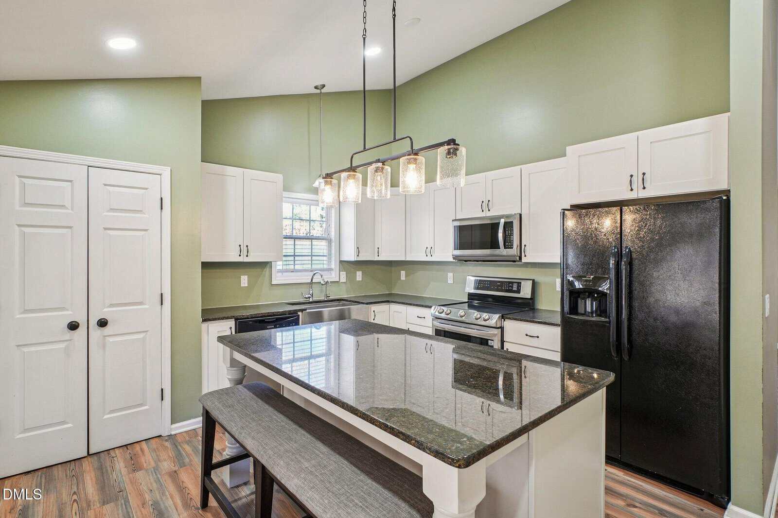 Modern kitchen with sage green walls, white cabinets, black appliances, a large island with a black countertop, a bench on one side, pendant lighting above, and wood-look flooring.