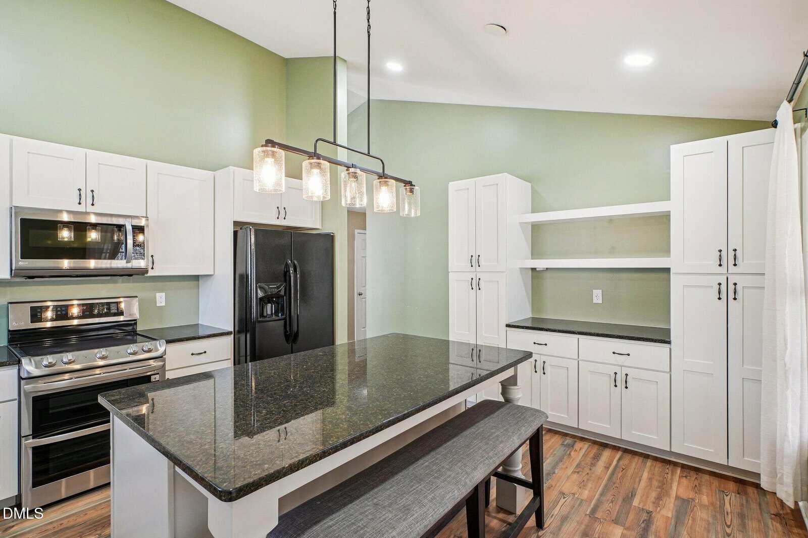Modern kitchen with white cabinets, black countertops, stainless steel appliances, a black refrigerator, a kitchen island with seating, pendant lights, open shelves, and light green walls with wood flooring.