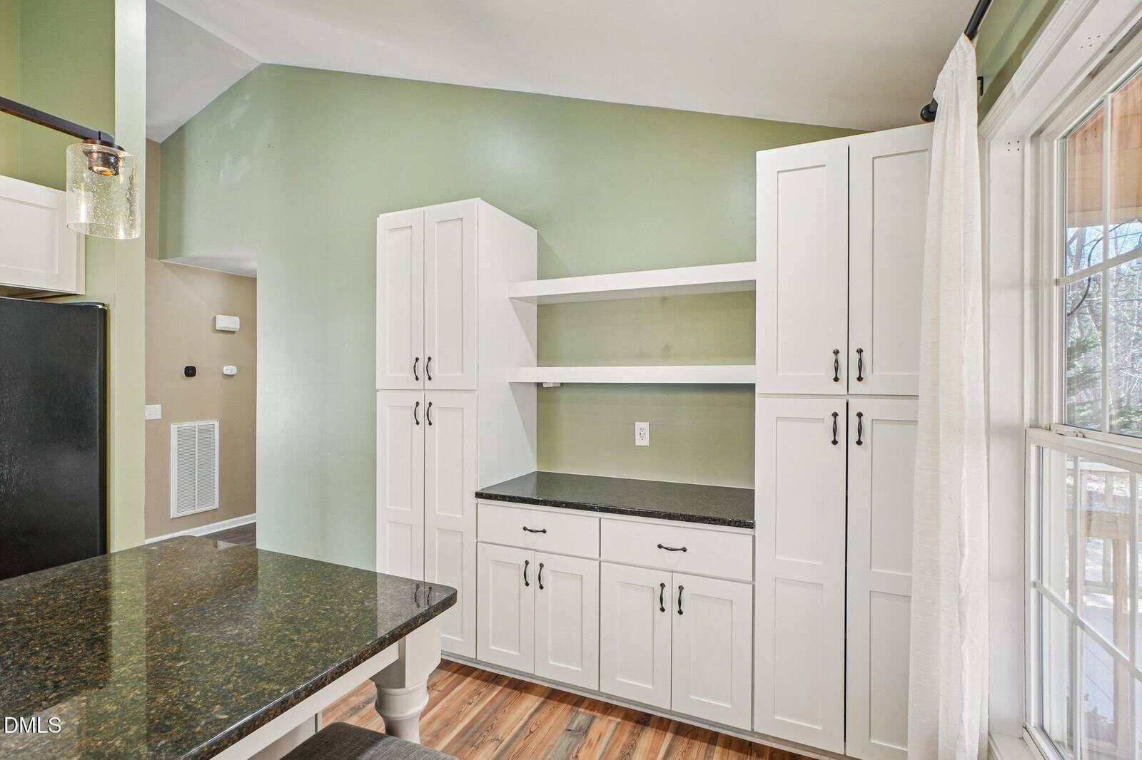 A kitchen with white cabinets, black countertops, light green walls, wood flooring, and large windows with white curtains letting in natural light.