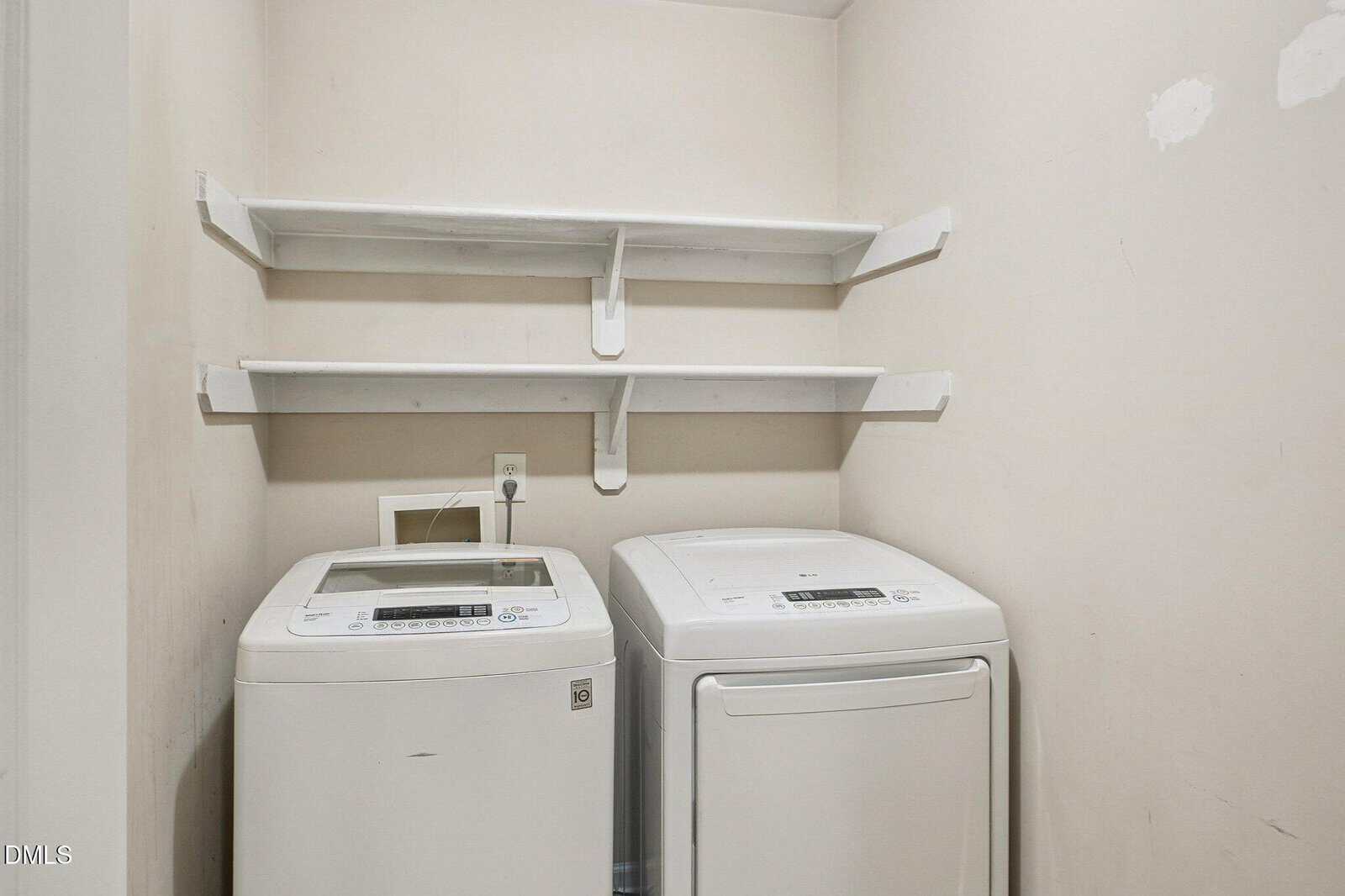 A small laundry room with a white top-loading washing machine and a white front-loading dryer beneath two white wall-mounted shelves on a beige wall.