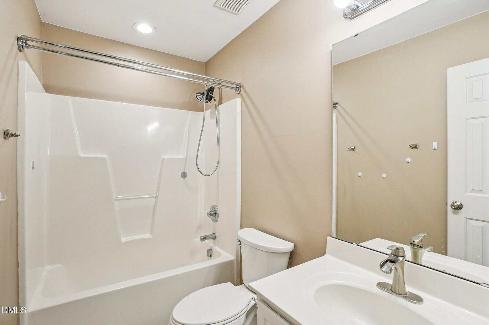 A bathroom with beige walls features a white bathtub-shower combo, toilet, and sink with a silver faucet. A large mirror hangs above the sink, reflecting the door and part of the room.