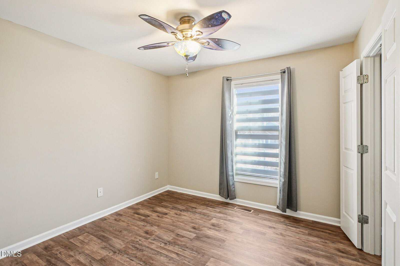 Empty room with light walls, wood-style flooring, ceiling fan with light, a window with sheer striped blinds and gray curtains, and a white double-door closet.