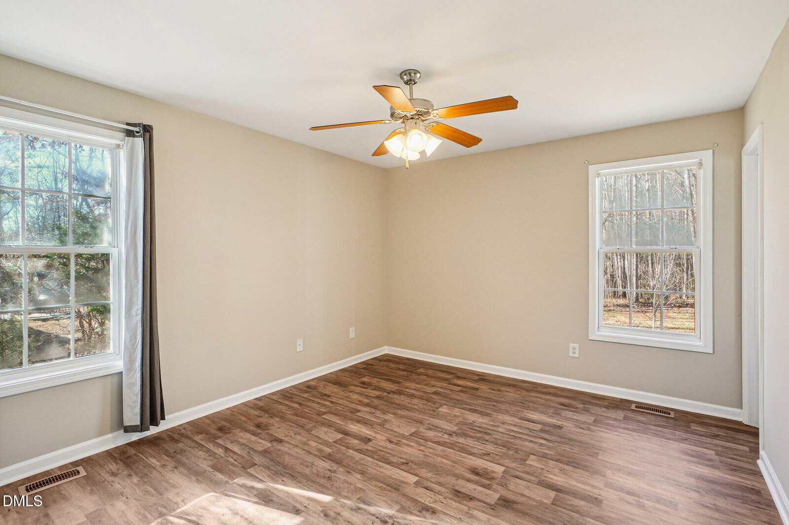 Bright empty room with beige walls, wood flooring, two large windows with white trim, and a ceiling fan with lights.