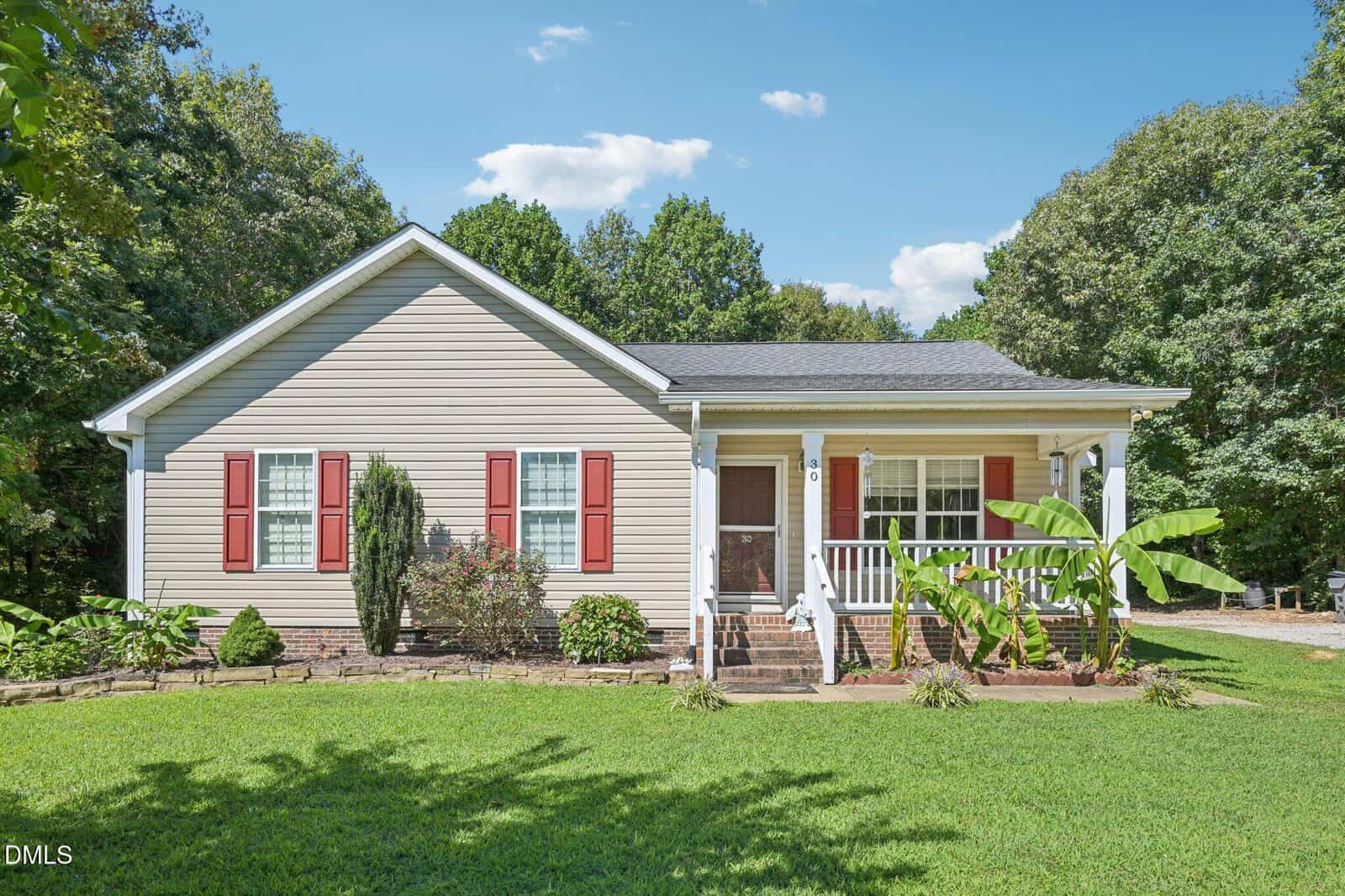 Single-story beige house with red shutters, a covered front porch, and white columns. The yard is neatly landscaped with green grass, shrubs, banana plants, and trees in the background under a blue sky with scattered clouds.