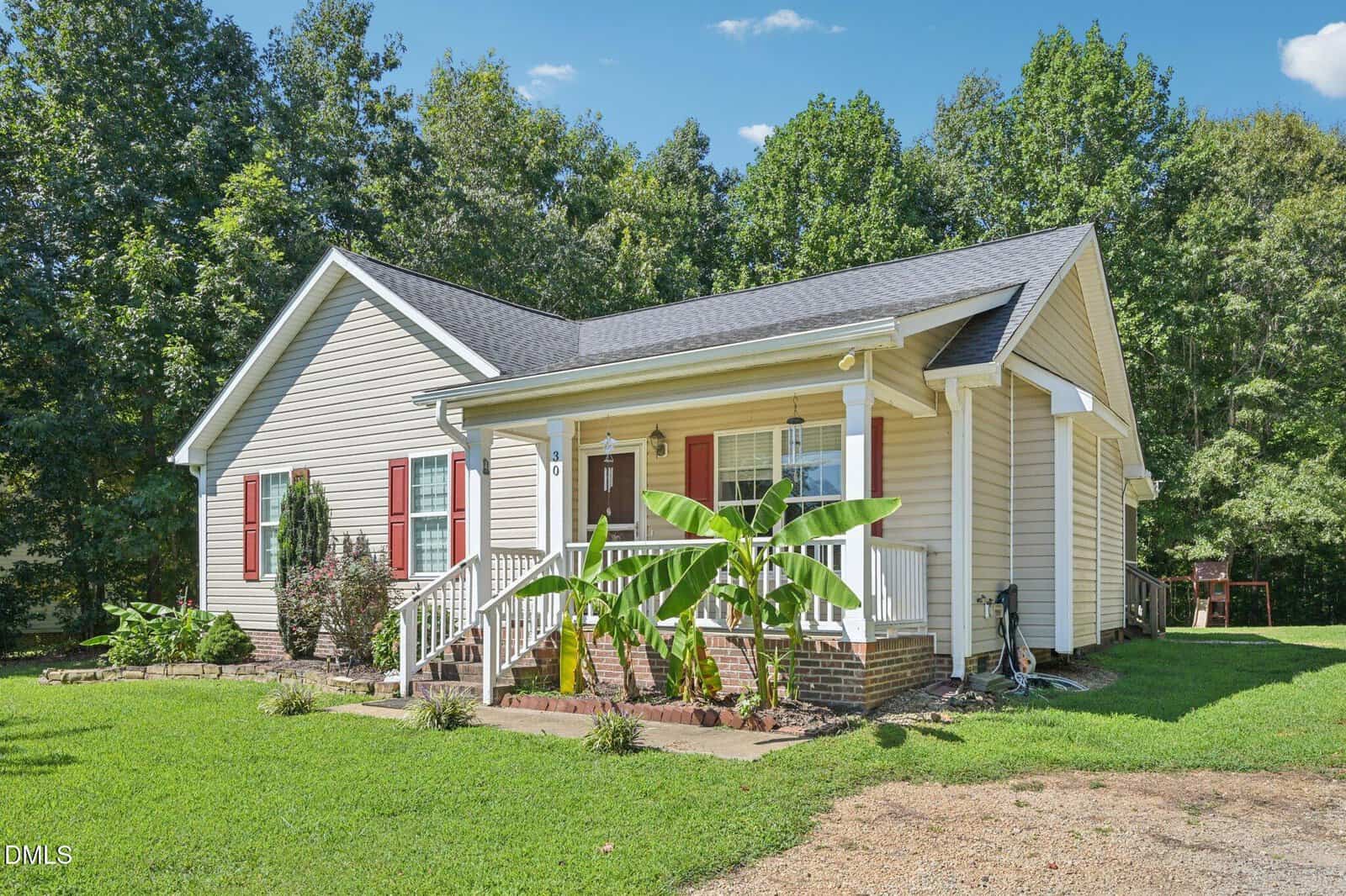Single-story beige house with a front porch, red shutters, and white railing. Green banana plants and shrubs are planted in front. The house is surrounded by a grassy yard and trees, with a clear blue sky above.