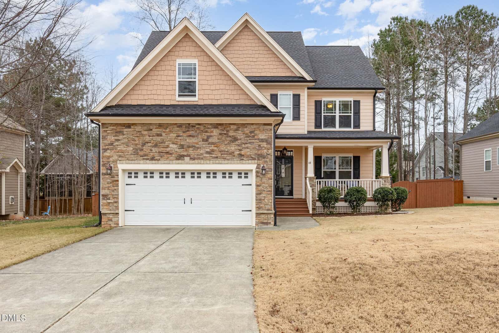 A two-story suburban house with tan siding, stone accents, a white double garage door, and a front porch. The yard has patchy brown grass and is bordered by trees and a wooden fence.