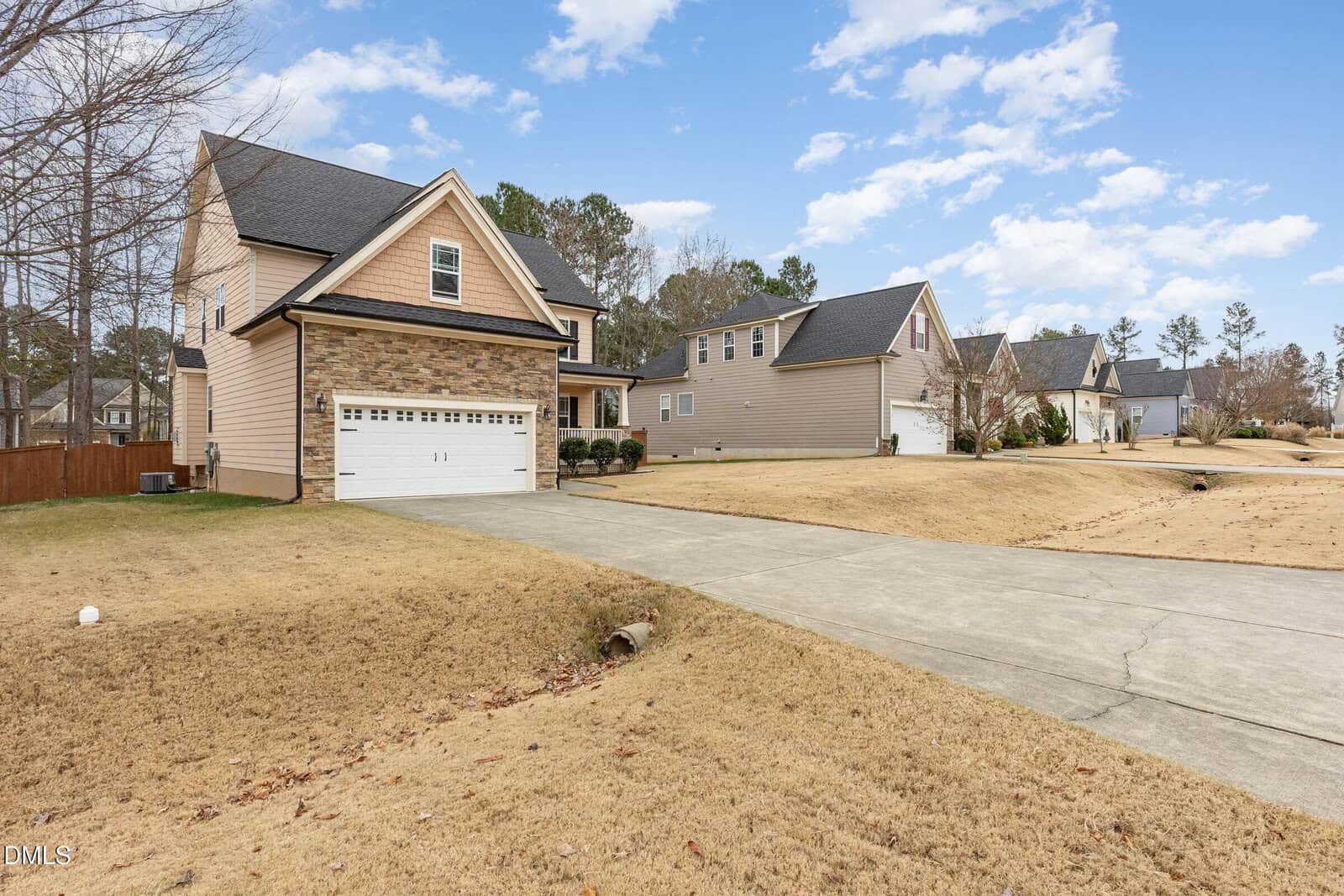 A suburban house with tan siding and stone accents, a two-car garage, and a wide driveway. Neighboring houses and trees line the street under a partly cloudy sky. The lawn is dry and brown.