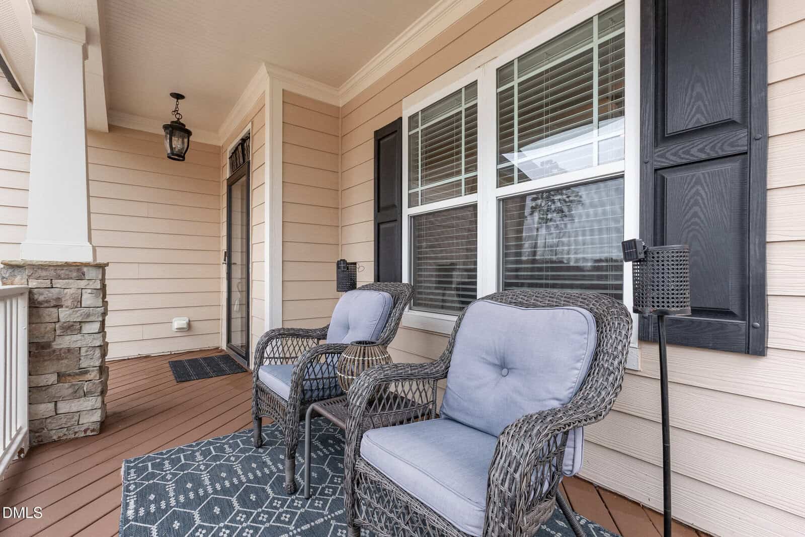 Two wicker chairs with blue cushions sit on a patterned rug on a covered porch with tan siding, black shutters, and a window, creating a cozy outdoor seating area.