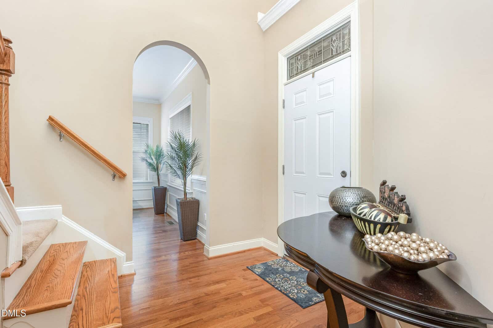 A bright entryway with hardwood floors, a white door, a decorative table with bowls, stairs with a wood railing, and two tall potted plants near a window in the adjoining room.