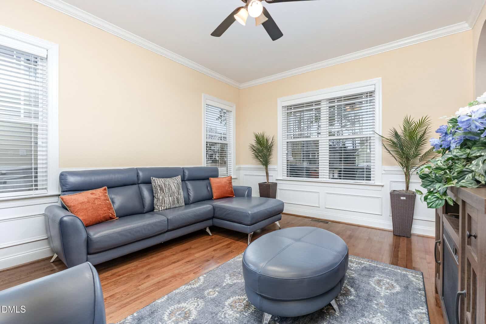 A bright living room with a blue sectional sofa, orange accent pillows, an oval ottoman, a blue patterned rug, potted plants near the windows, and a ceiling fan above. The room has wood floors and cream-colored walls.