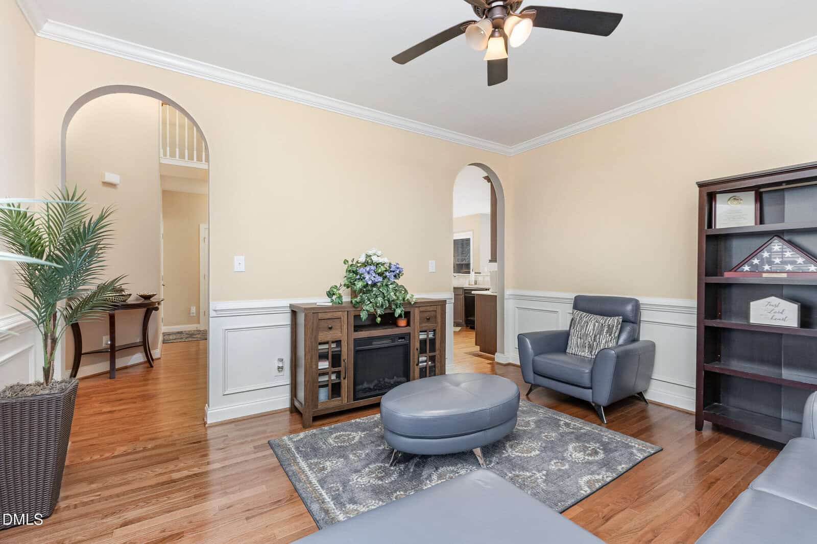 A bright living room with tan walls, white trim, wood floors, gray armchairs, a round ottoman, an electric fireplace with floral decor, a bookcase, a ceiling fan, and arched doorways leading to other rooms.