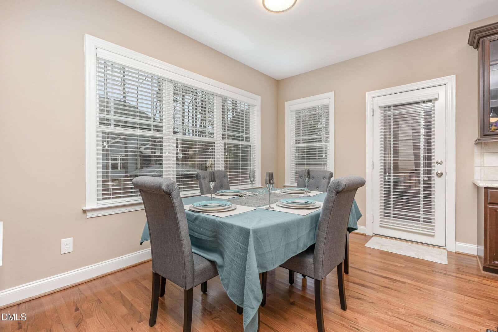 A dining area with a square table covered in a teal tablecloth, set for four with plates and glasses. Four gray upholstered chairs surround the table. Large windows and a glass door let in natural light. The floor is wood.