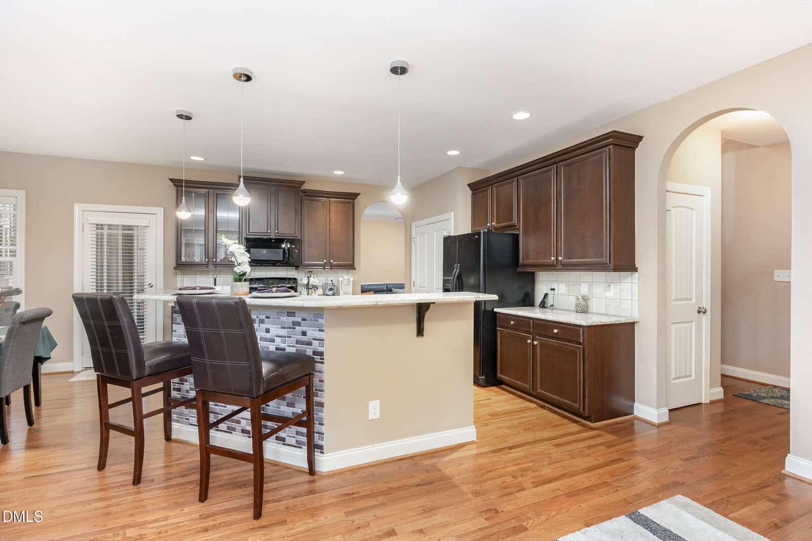 Modern kitchen with dark wood cabinets, black appliances, a breakfast bar with two brown chairs, pendant lights, white countertops, and hardwood flooring. An arched doorway and dining area are visible in the background.