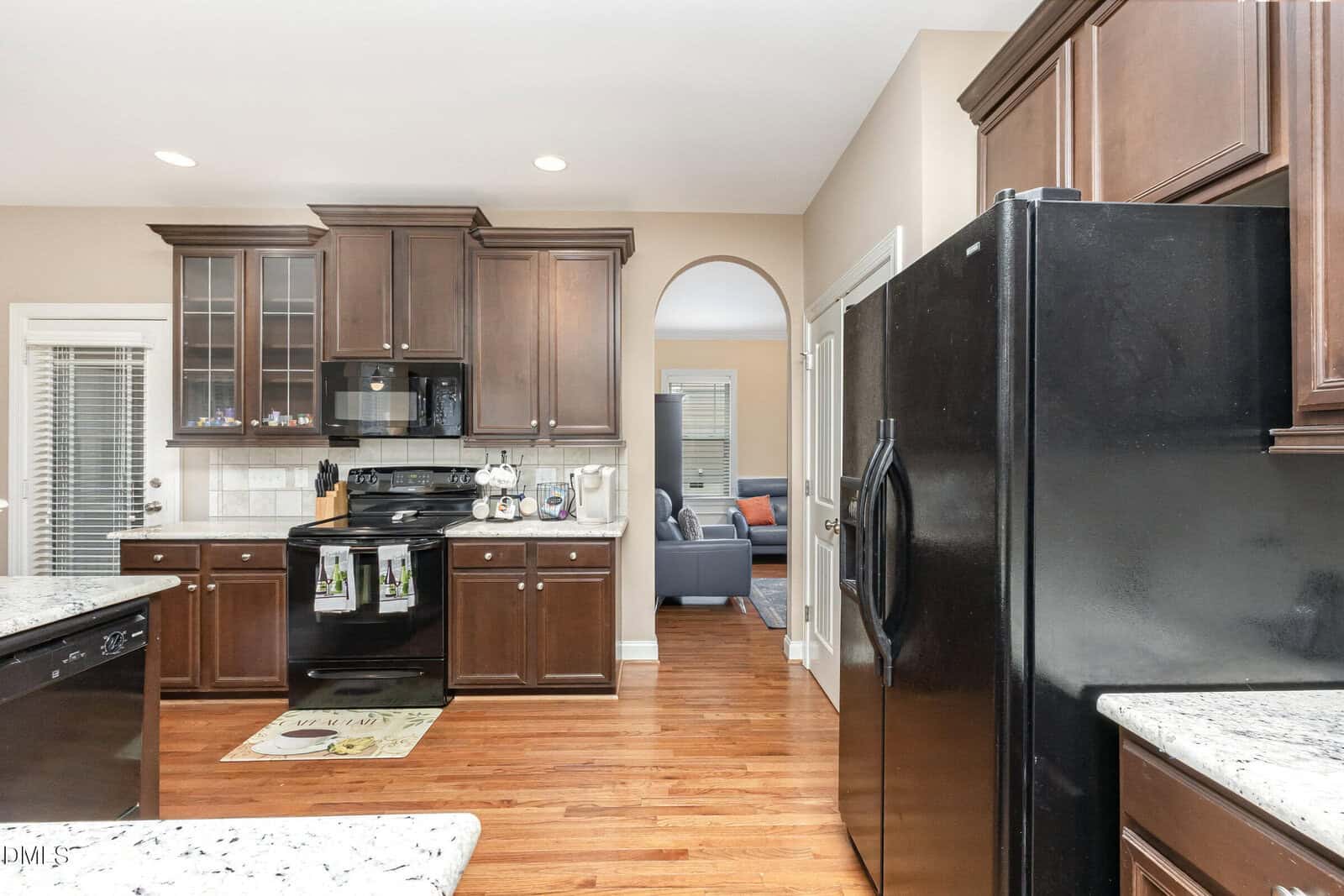 A modern kitchen with dark wood cabinets, black appliances, and white granite countertops. Hardwood floors and an arched doorway lead to a living area with a gray sofa in the background.