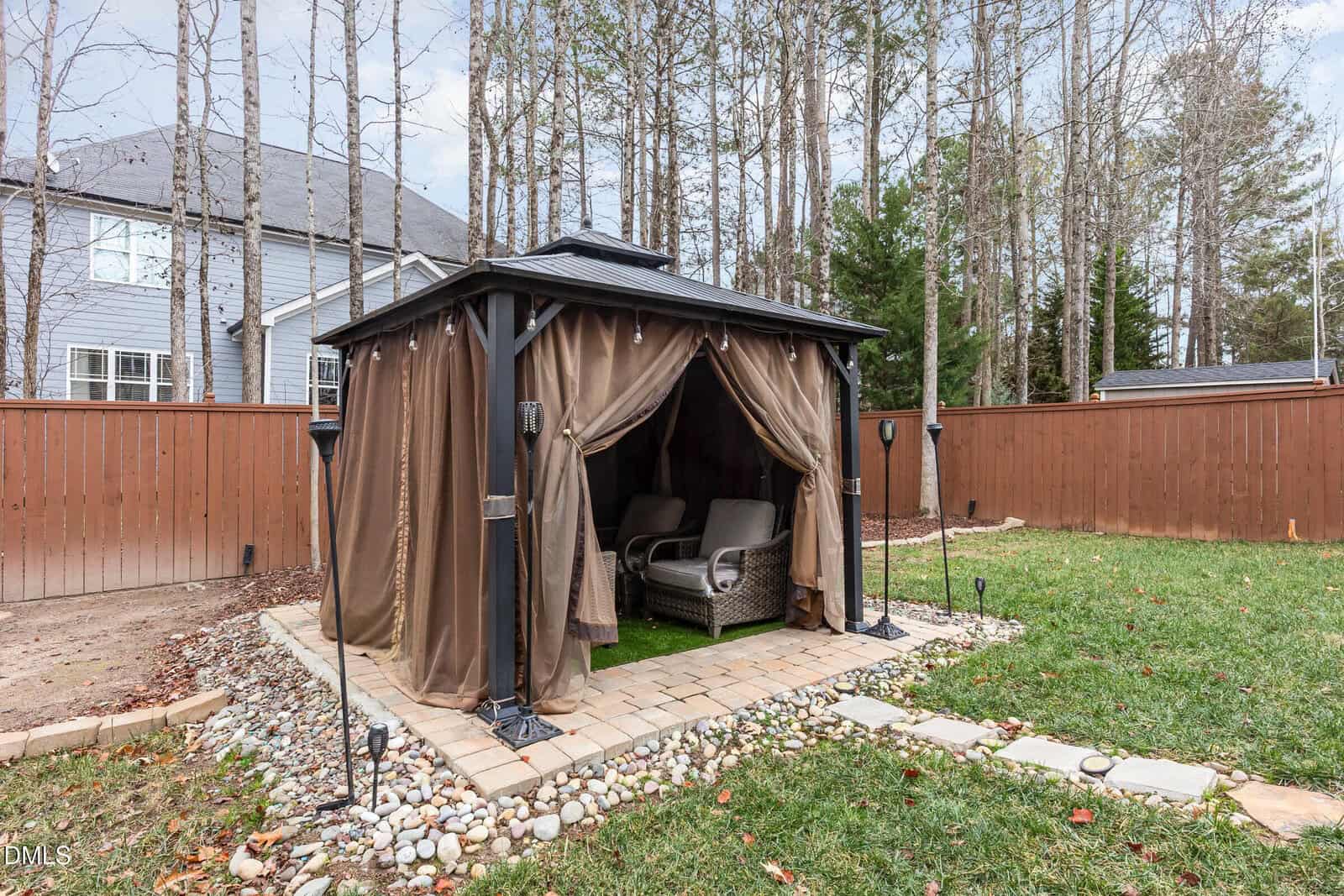 A backyard gazebo with brown curtains, two cushioned chairs inside, and a stone pathway, surrounded by grass, trees, and a wooden fence.
