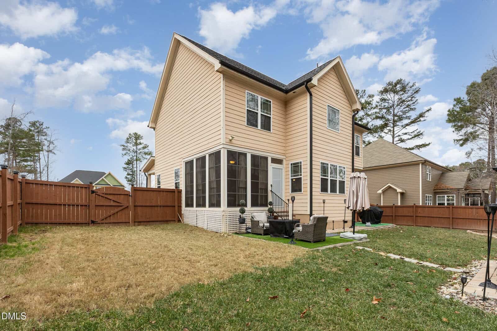 Two-story beige house with a screened-in porch, outdoor seating area, fenced backyard, and patio umbrella. The yard has patchy grass and is surrounded by several neighboring houses and tall trees under a partly cloudy sky.