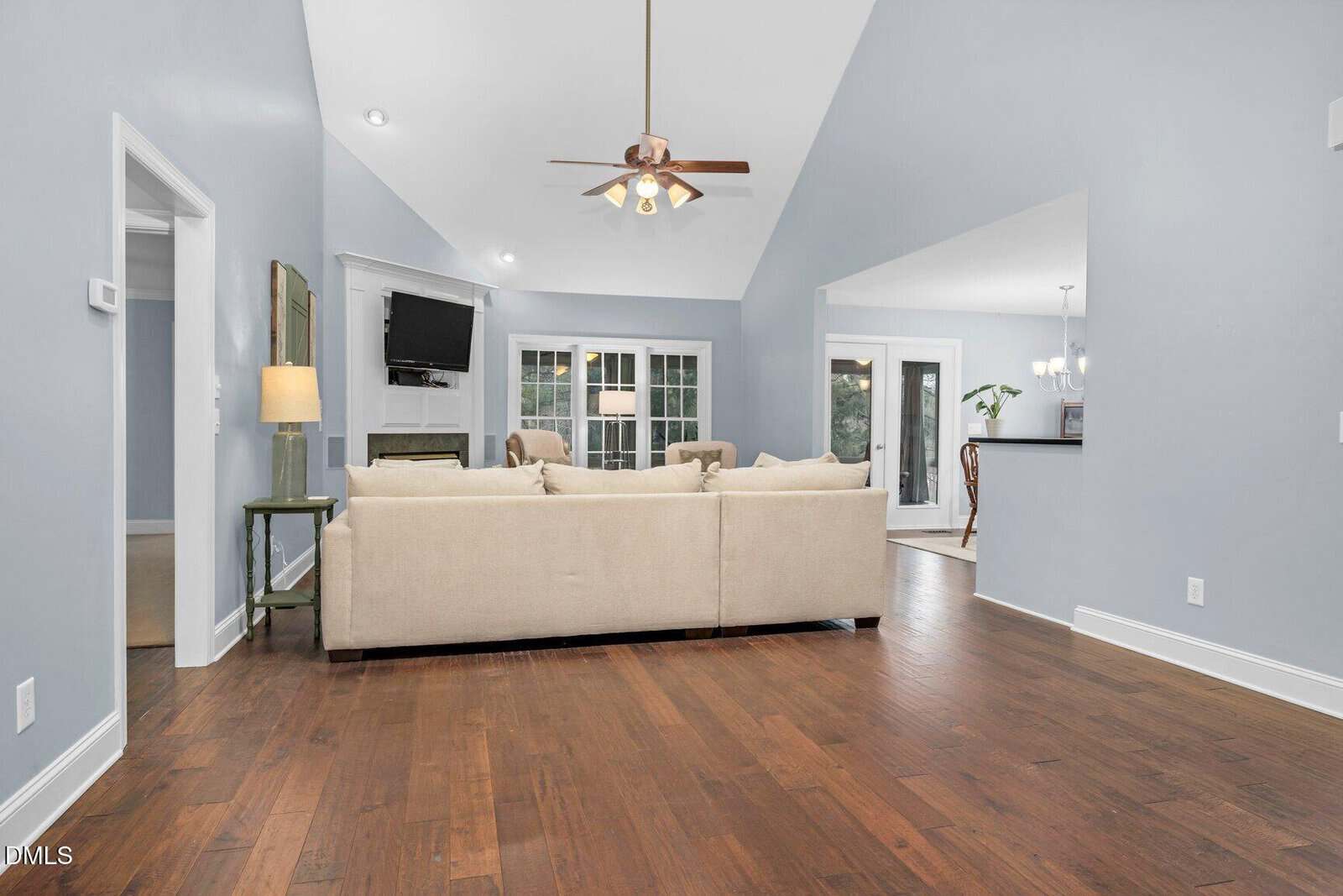 A spacious living room with light blue walls, dark hardwood floors, a beige sectional sofa, ceiling fan, wall-mounted TV above a fireplace, and large windows leading to a bright dining area.