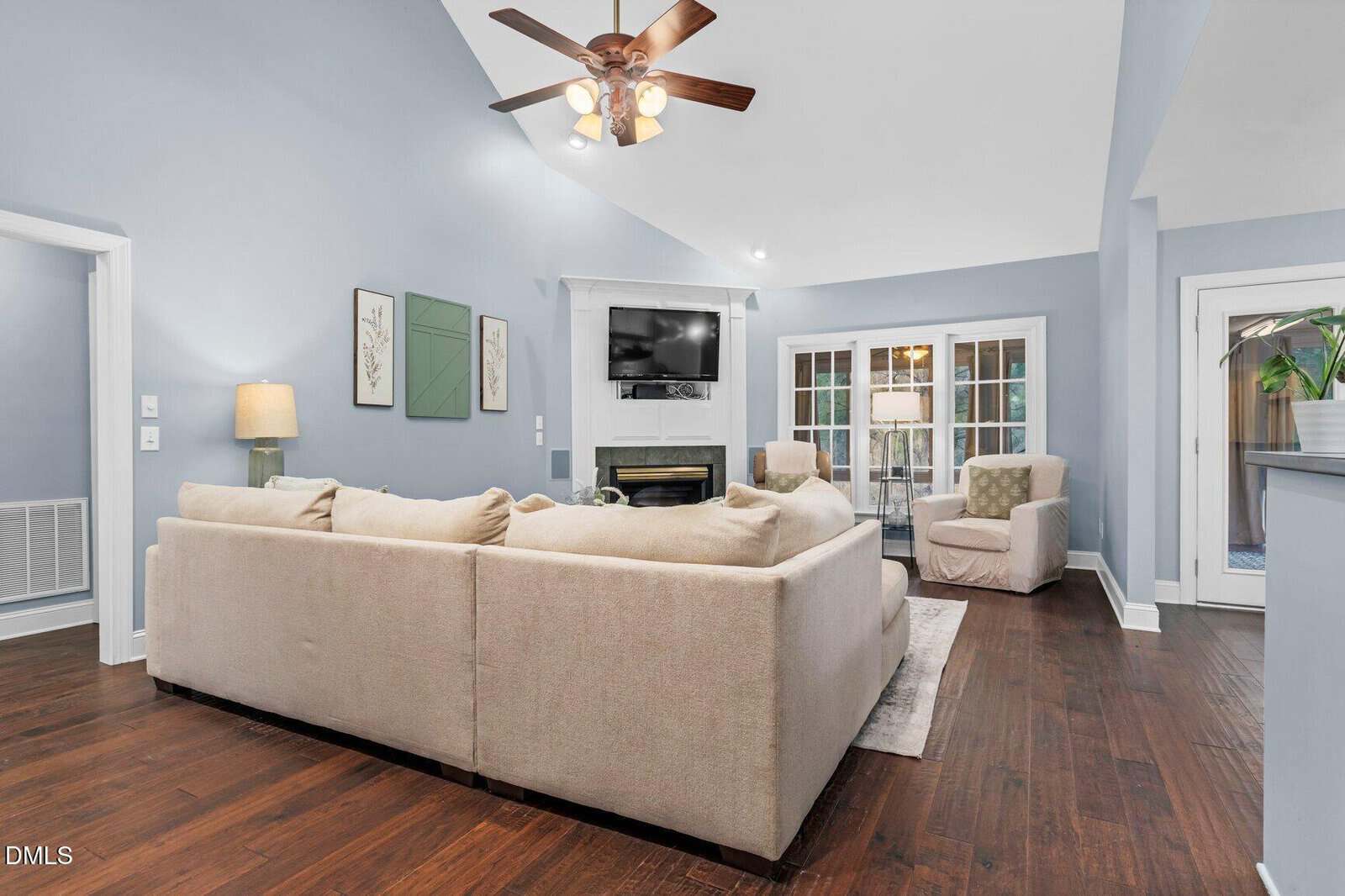 Spacious living room with light blue walls, a beige sectional sofa, armchair, ceiling fan, wall-mounted TV above a fireplace, hardwood floors, and glass doors leading to a patio.