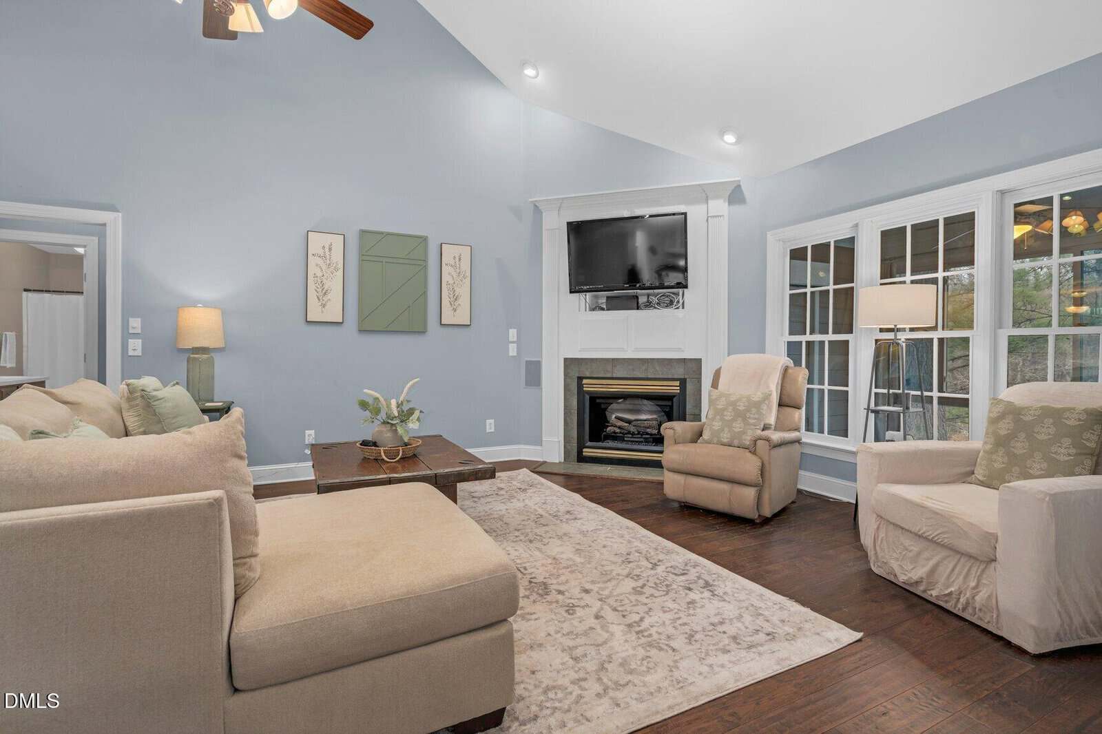 A cozy living room with light blue walls, beige sectional sofa, two beige armchairs, a fireplace with a TV above it, a large window, and hardwood floors. Decorative art and lamps add warmth to the space.
