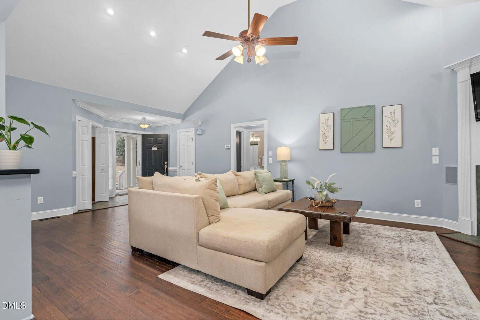 A bright living room with light blue walls, vaulted ceiling, and a ceiling fan. There is a beige sectional sofa, a wooden coffee table, a rug, and wall art. The entryway and plants are visible in the background.