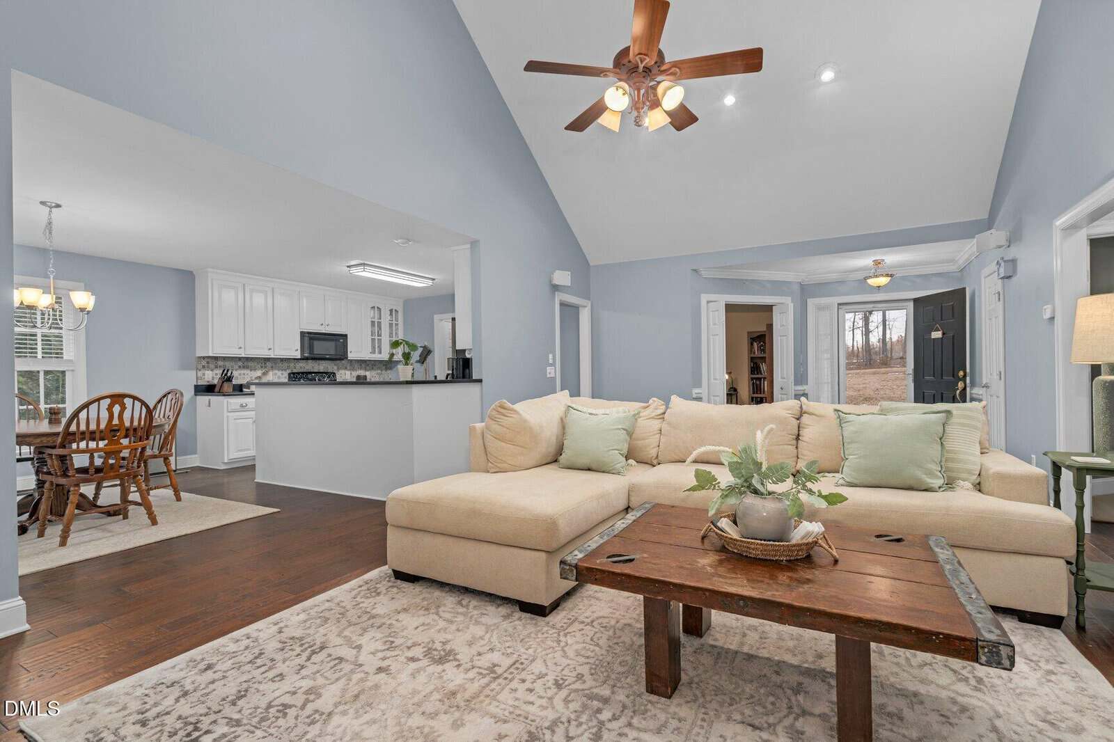 Open living room with a beige sectional sofa, wooden coffee table, light patterned rug, and ceiling fan; adjacent to a kitchen with white cabinets and a dining area with a wooden table and chairs.
