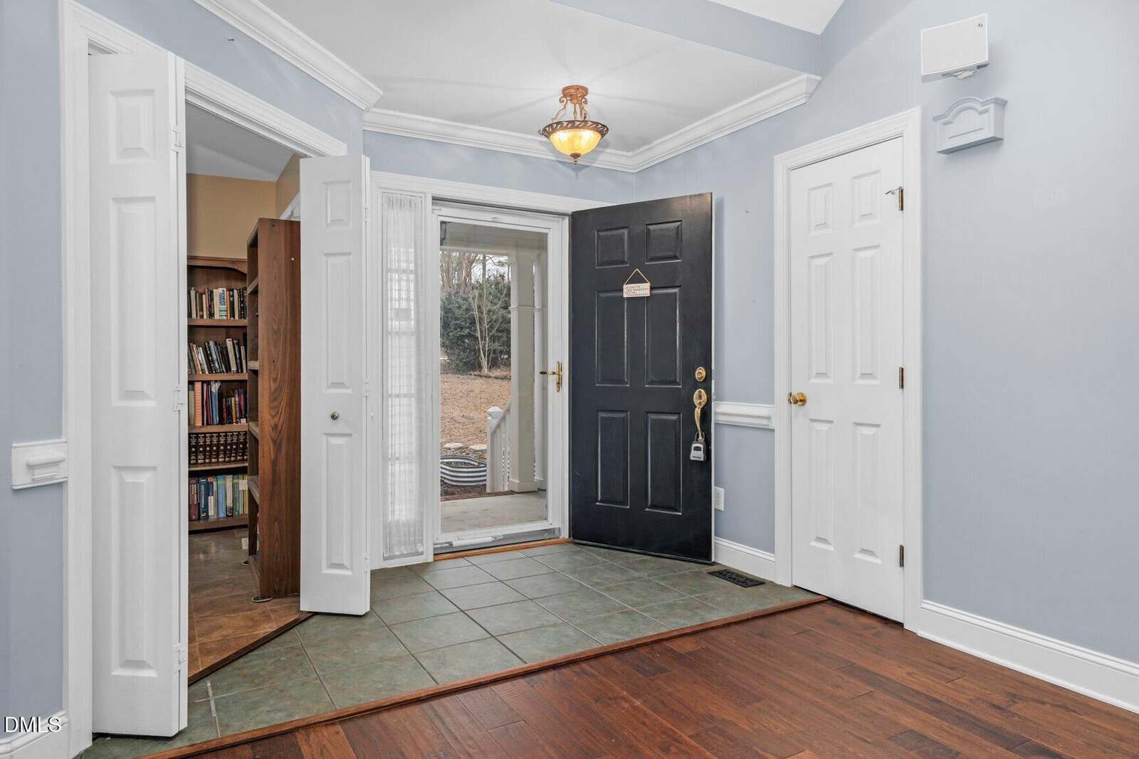 A home entryway with a black front door open to the outside, white double doors revealing a room with bookshelves, blue walls, and a mix of tile and wood flooring under a ceiling light.