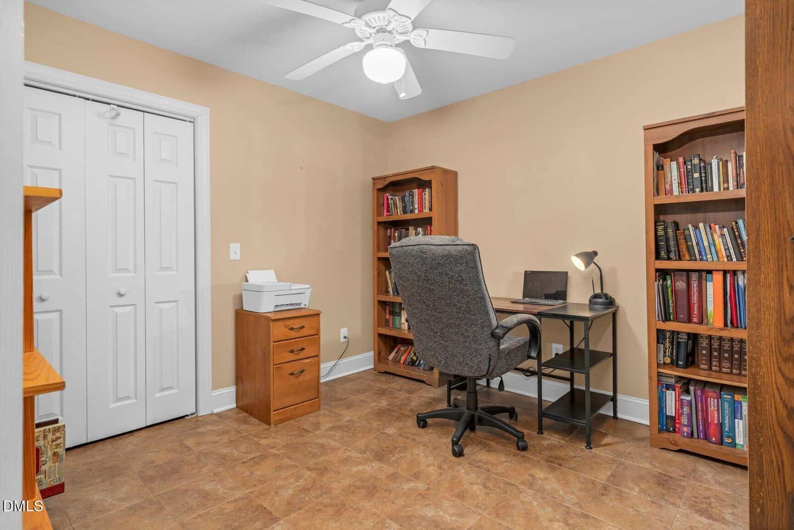 A home office with tan walls, tile flooring, a ceiling fan, a desk with a lamp and laptop, an office chair, a wooden filing cabinet with a printer, and two bookshelves filled with books.