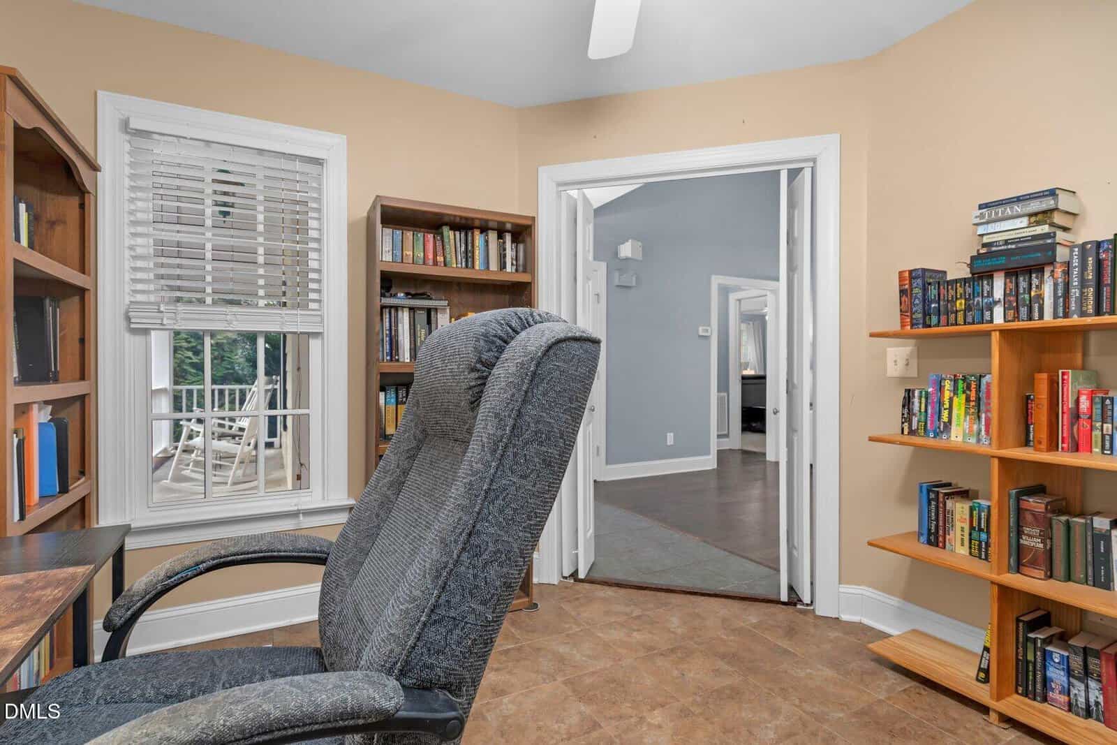 A home office with a gray office chair, wooden desk, bookshelves filled with books, tile floor, and a window with blinds. Double doors open to a hallway with blue walls and wooden flooring.