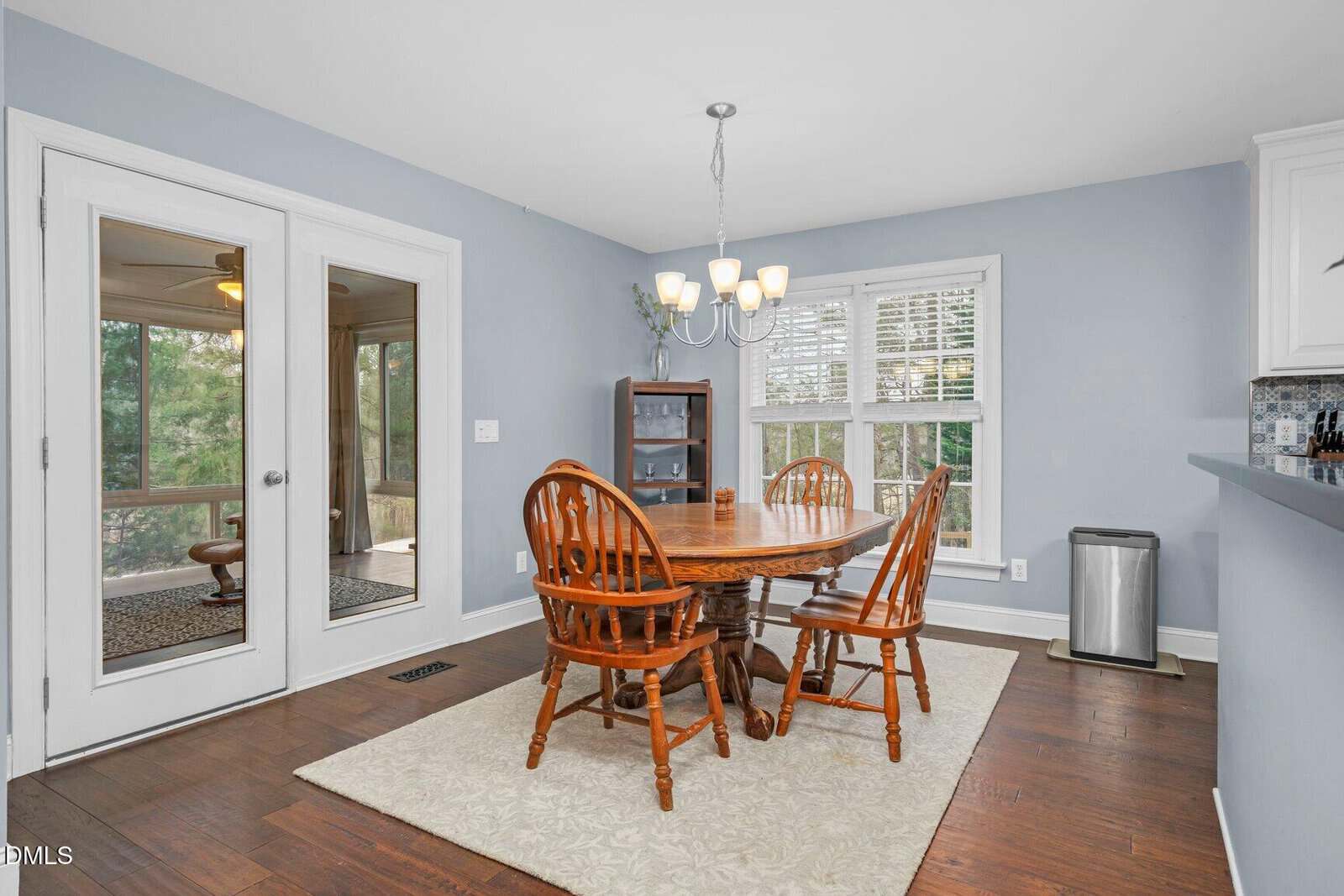 Bright dining area with a round wooden table, four chairs, a light fixture, hardwood floors, light blue walls, large windows with blinds, a small cabinet, and double glass doors leading to an outdoor space.