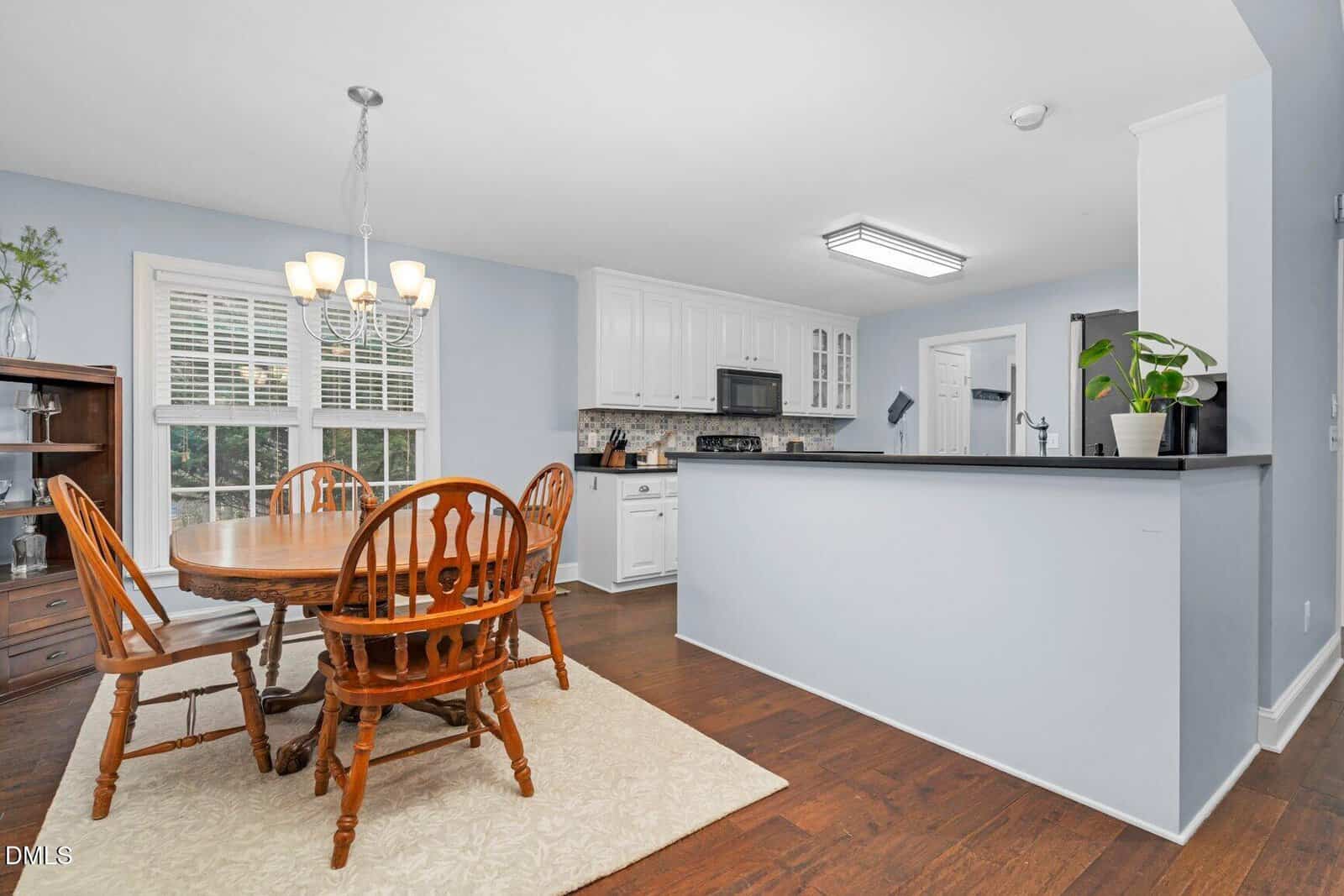 A kitchen and dining area with wooden table and chairs, white cabinets, dark countertops, and hardwood floors. A window with blinds, a light fixture, and houseplants add brightness to the space.