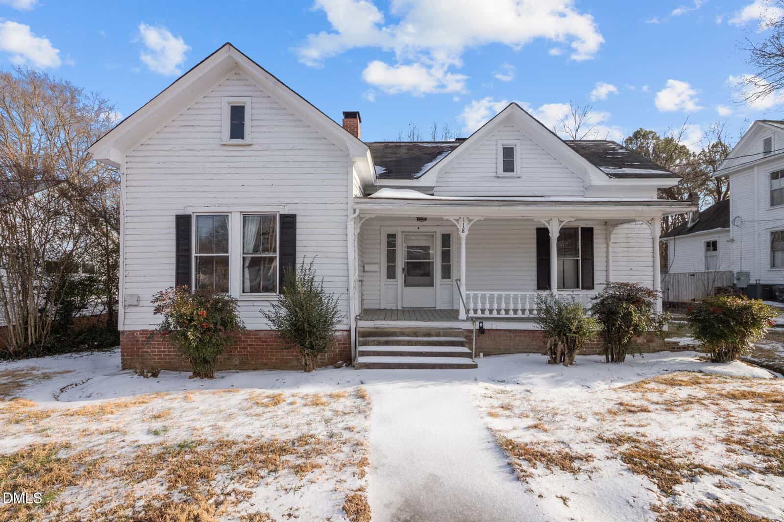 A white wooden house with dark shutters, a covered front porch, and steps leading to the entrance. The yard is partially covered with snow, and some bushes are near the porch. The sky is blue with scattered clouds.